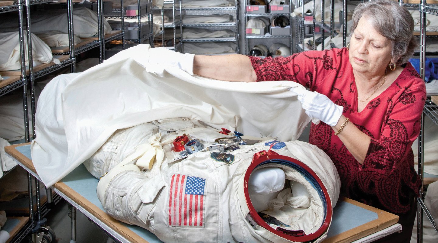 A woman wearing gloves covers a used astronaut suit with a sheet to preserve it.